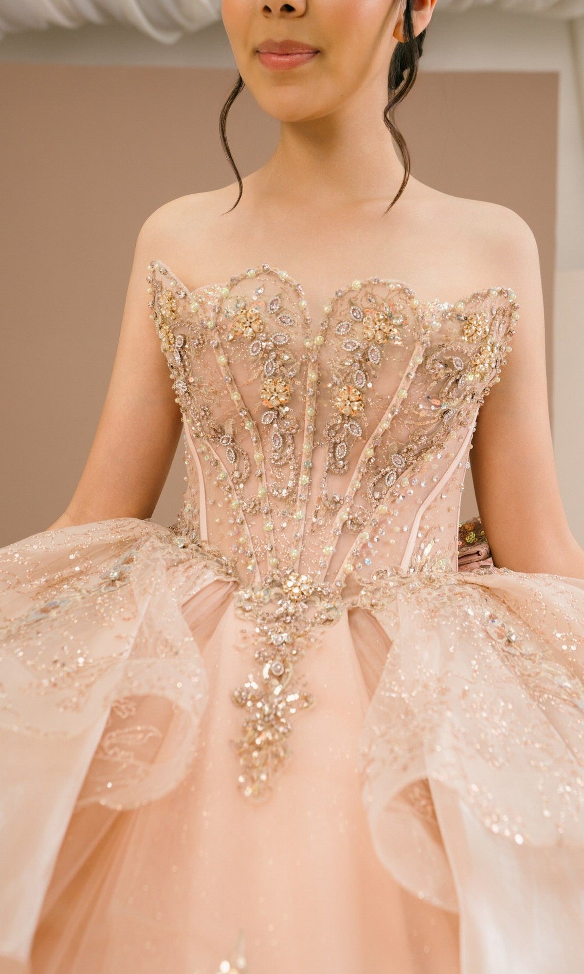 A woman wearing the Quinceanera Dress 8590JQ by Cinderella Couture, a strapless peach ball gown with a beaded corset bodice featuring intricate gold and silver beadwork, and a glitter tulle skirt adorned with sparkling embellishments.